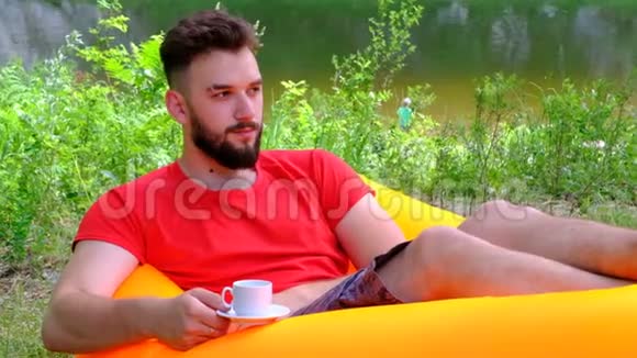一个穿着红色T恤和一杯咖啡的年轻人躺在河底的通风的沙发上那个家伙视频的预览图