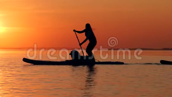 日落背景下的女人和狗划桨视频的预览图