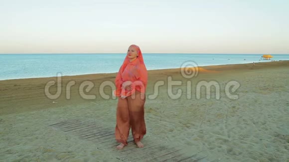 一位身穿橙色围巾的年轻穆斯林妇女站在海边站在大海的背景下视频的预览图