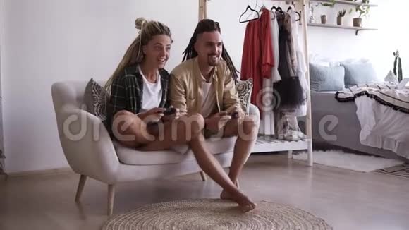 可爱的年轻夫妇男女朋友正在玩电子游戏手里拿着无线操纵杆坐在白色的小东西上视频的预览图