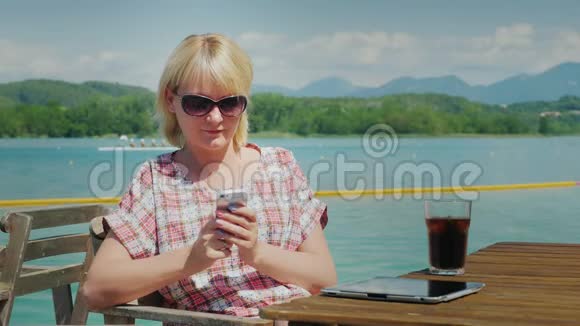 商务女性在度假时使用智能手机坐在湖边的一家咖啡馆里的一张桌子上背景是群山视频的预览图