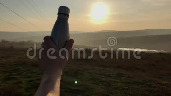 在日落背景下用男性手握钢制生态热瓶进行水的特写视频的预览图