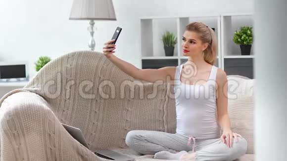 迷人的微笑年轻女孩放松坐在沙发上在家里用智能手机自拍视频的预览图