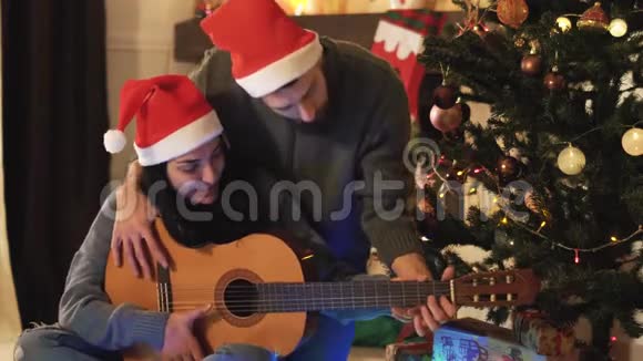 圣诞前夜圣诞老人戴帽子的幸福夫妇的肖像男人教女人弹吉他圣诞树上的圣诞树视频的预览图