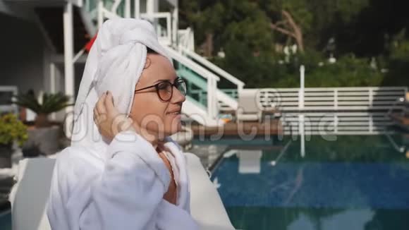 戴着眼镜穿着白色长袍头上拧着毛巾的女人正坐在露天的泳池边享受着视频的预览图