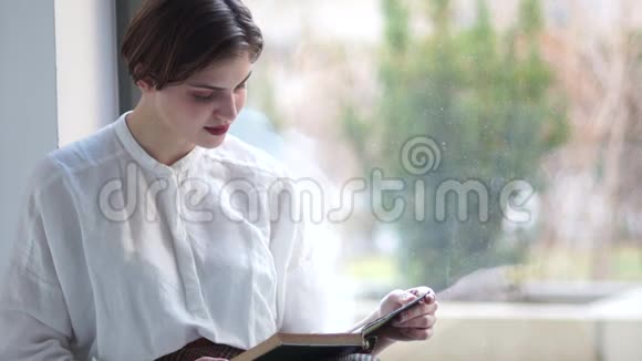 女人坐在窗户前看书一个穿着一件老式白色衬衫的年轻女孩的近距离室内画像视频的预览图