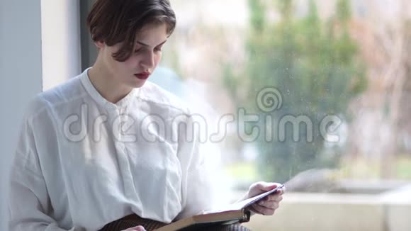 穿着白色衬衫的悲伤女孩坐在窗户上手里拿着一本书女学生度假快乐的女人视频的预览图