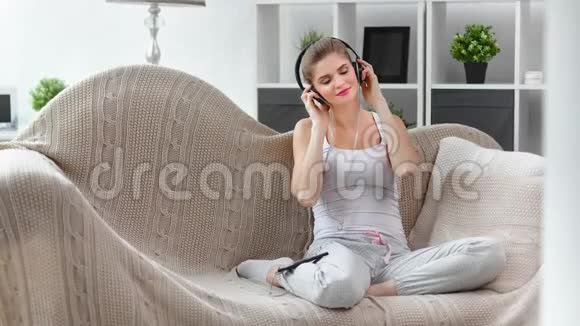 可爱的年轻女性穿着智能手机听着耳机在沙发上放松视频的预览图