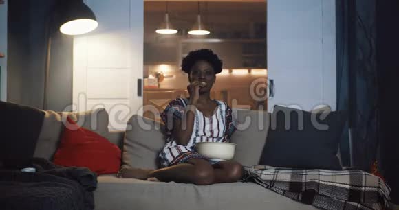 快乐的年轻休闲非洲女人坐在沙发上在家里看电影放映机与饮料和爆米花慢动作视频的预览图