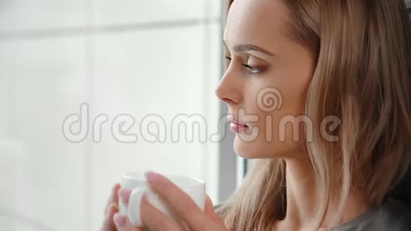 从杯子里看着窗外一个安静的年轻女人喝着热饮料的特写视频的预览图