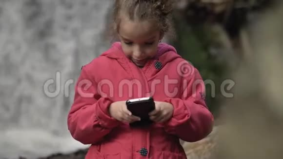5岁的小女孩在美丽的大自然中站在瀑布前用智能手机视频的预览图