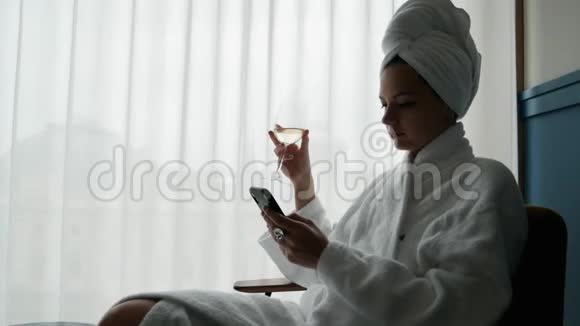 优雅的女人穿着白色浴袍头上有一条毛巾享受着一杯葡萄酒在酒店里使用手机视频的预览图