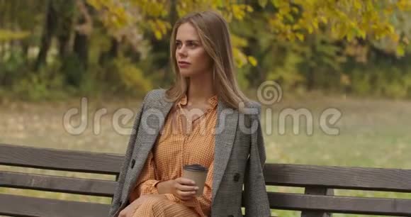 美丽的高加索女人坐在秋天公园的长凳上喝着一杯咖啡漂亮的姑娘摇着她视频的预览图