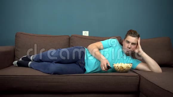 一个年轻人正在看电视和吃爆米花那家伙躺在沙发上从电视上看到的我看着视频的预览图