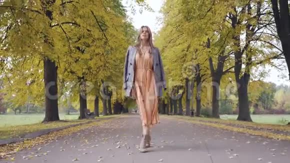 穿着芥末色连衣裙和一件格子夹克的白种人黑黑肤色少女在铺满黄叶的道路上行走视频的预览图