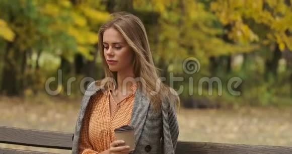 一个可爱的高加索女人坐在秋天公园的长凳上喝咖啡的肖像可爱的女孩视频的预览图