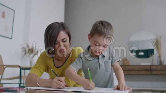 母亲穿着黄色夹克儿子穿着t恤坐在桌子旁在纸上画着彩色铅笔快乐的童年视频的预览图