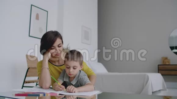 母亲穿着黄色夹克儿子穿着t恤坐在桌子旁在纸上画着彩色铅笔快乐的童年视频的预览图