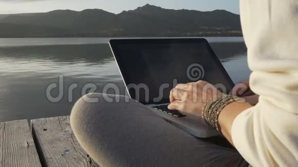 互联网自由职业自由职业自由职业选择概念一位年轻女性在日落时分坐在湖边的码头上的笔记本电脑上工作视频的预览图