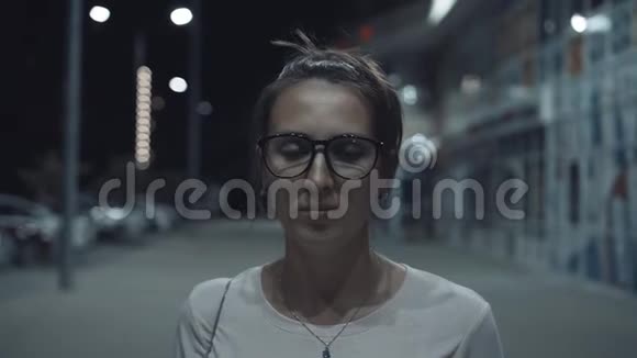 一个戴着眼镜的快乐帅气的女孩看着镜框微笑着坐落在城市的夜街上视频的预览图