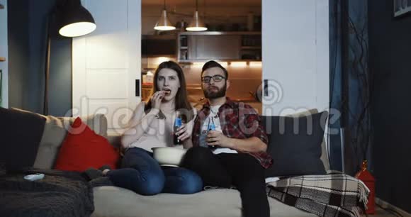 快乐年轻开朗的欧洲男人和女人坐在一起在家里看有趣的电影带着零食慢动作视频的预览图