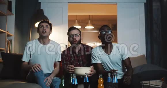开朗多民族的年轻男球迷喊着一起在家看足球运动比赛带着零食慢动作视频的预览图