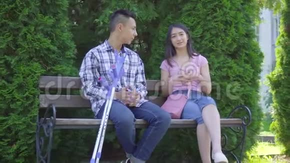 年轻积极的亚裔男子拄着拐杖断腿与坐在长凳上的年轻亚裔女子交流视频的预览图