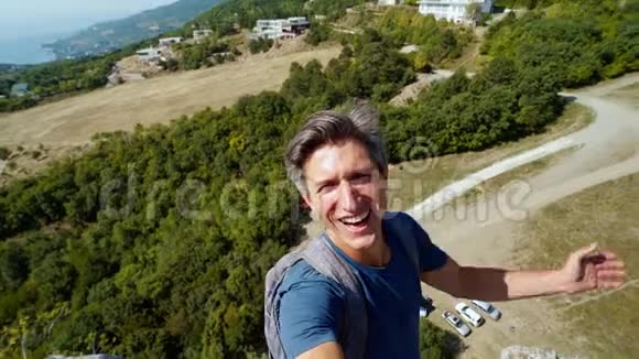 微笑的男人摄影师捕捉视频自拍站在悬崖边的山天空和海洋背景视频的预览图