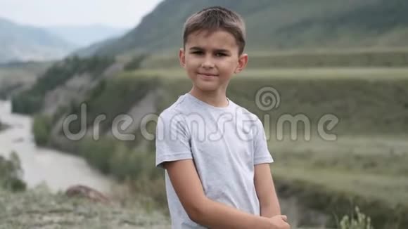 肖像一个穿着灰色T恤的小旅游男孩看着山和河背景上的摄像机视频的预览图