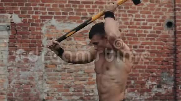 强壮的肌肉赤膊的年轻人用大锤打轮胎背景上的红砖楼关门视频的预览图