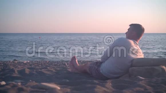 一个年轻人坐在岸边映衬着海上日落日落时分人们正坐在海滩上视频的预览图