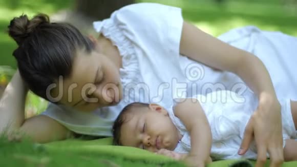 妈妈吻她的孩子睡在绿草的户外绿色夏日公园里微笑的年轻母亲和孩子视频的预览图