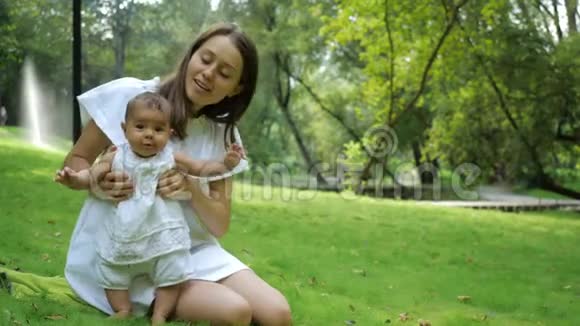 年轻漂亮的母亲穿着一件白色的衣服教她刚出生的女儿在公园里的一片新鲜的绿草上行走年轻快乐视频的预览图