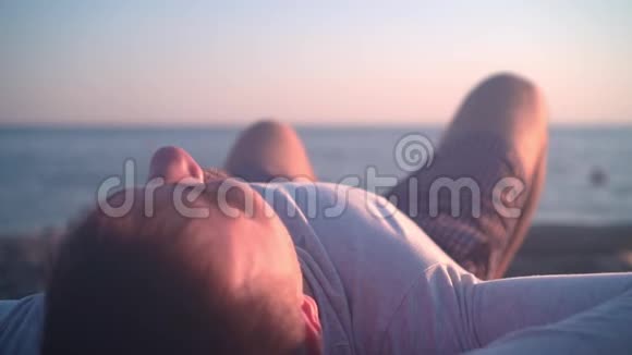 一个年轻人躺在海岸上背景是海上日落特写镜头在日落的背景下躺在海滩上视频的预览图