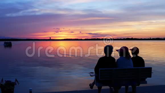 三位资深女性在一个美丽的湖上坐在长凳上欣赏五颜六色的日落视频的预览图