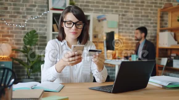 年轻妇女在工作中使用信用卡和智能手机在线支付视频的预览图