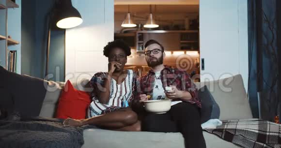 快乐的年轻休闲高加索男人和非洲女人在家看电视剧聊天吃爆米花慢动作视频的预览图