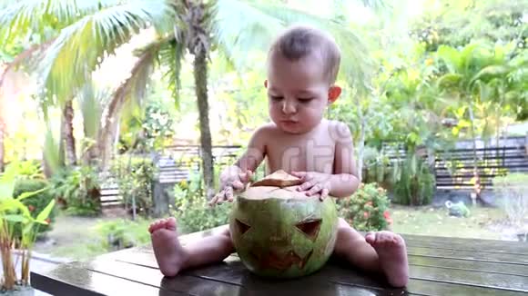 一岁的婴儿坐在棕榈树背景下的木桌上拥抱着一个绿色的新鲜椰子一张脸可以视频的预览图