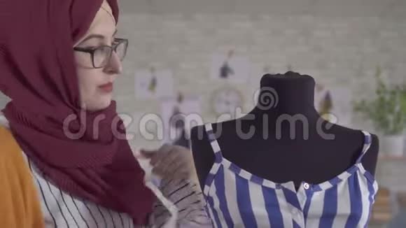 年轻的穆斯林女时装设计师戴着国家头巾在一个人体模特身上做一件蓝色连衣裙视频的预览图
