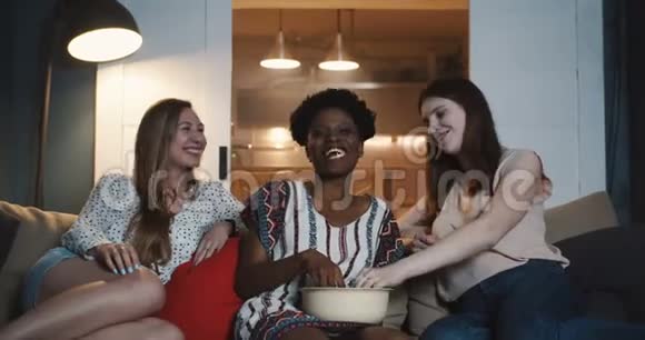 三个快乐的年轻多民族女性表达不同的情感在家里用零食慢动作看电影视频的预览图