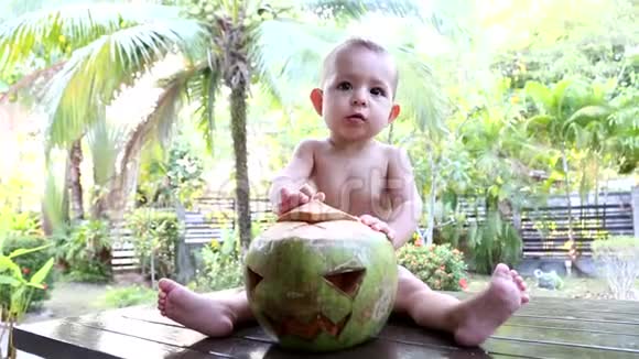 婴儿坐在一张木桌上背景是棕榈树和有趣的哈欠在他面前是一个绿椰子视频的预览图