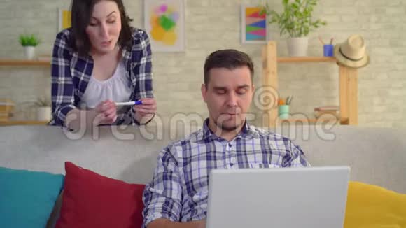 年轻积极的妻子高兴地跳舞她怀孕了悲伤的丈夫震惊地坐在笔记本电脑前的沙发上视频的预览图