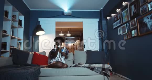 快乐年轻开朗的黑人丈夫和妻子一起坐在家里的沙发上看电影带着零食慢动作视频的预览图