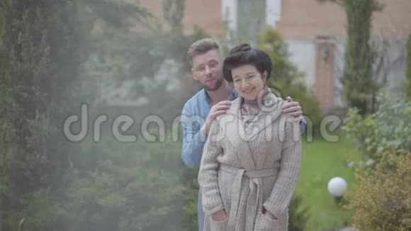 画像上幸福的成年女子站在花园里的大房子前面成年的孙子抱着她放着视频的预览图