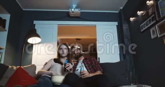 快乐的年轻白种人新婚家庭坐在一起看电影在家里与饮料使用投影仪慢动作视频的预览图