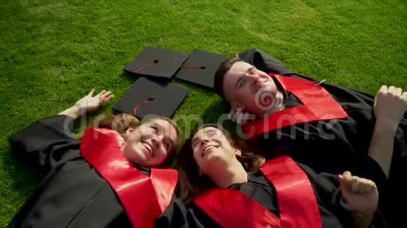 三个快乐的微笑学生穿着黑色的斗篷躺在草地上享受她的自由年轻人庆祝他们视频的预览图