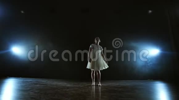 一个穿着白色连衣裙的女孩跳着现代芭蕾舞在舞台上用聚光灯下的烟雾旋转视频的预览图