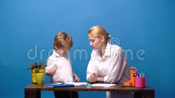 母亲坐在家里的桌子上帮她的小儿子做蓝色背景的家庭作业女人和小孩视频的预览图