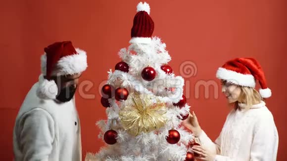 男人和女人用红色背景上的红色球装饰白色圣诞树爱的情侣准备新年晚会视频的预览图