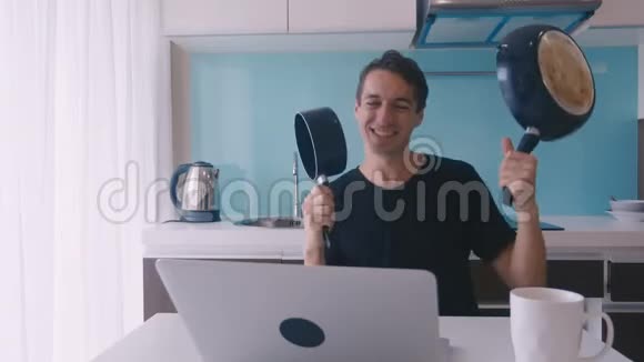 疯狂的滑稽男子正拿着煎锅和平底锅跳舞坐在厨房的笔记本电脑旁庆祝视频的预览图